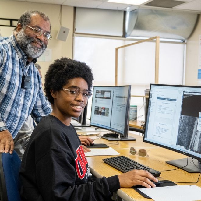 Student and instructor smiling at camera as they work at a computer station