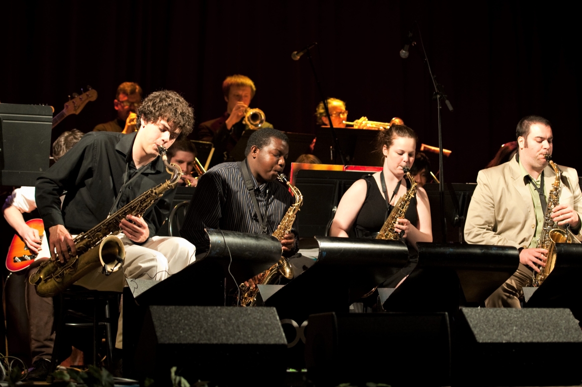 Students wearing black on stage in rows performing jazz music, playing saxaphones, trumpets, guitar and more.