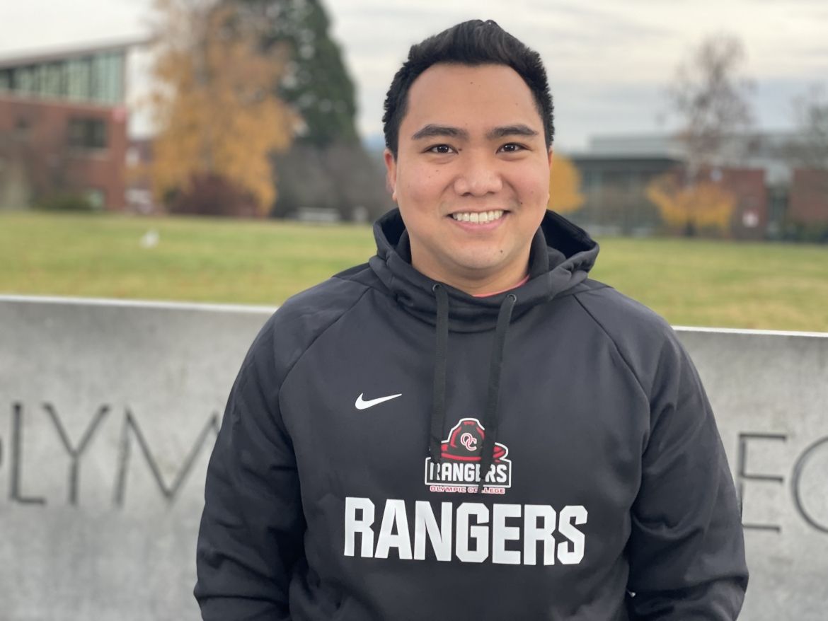 Smiling student in dark gray Rangers sweatshirt poses in front of cement Olympic College sign