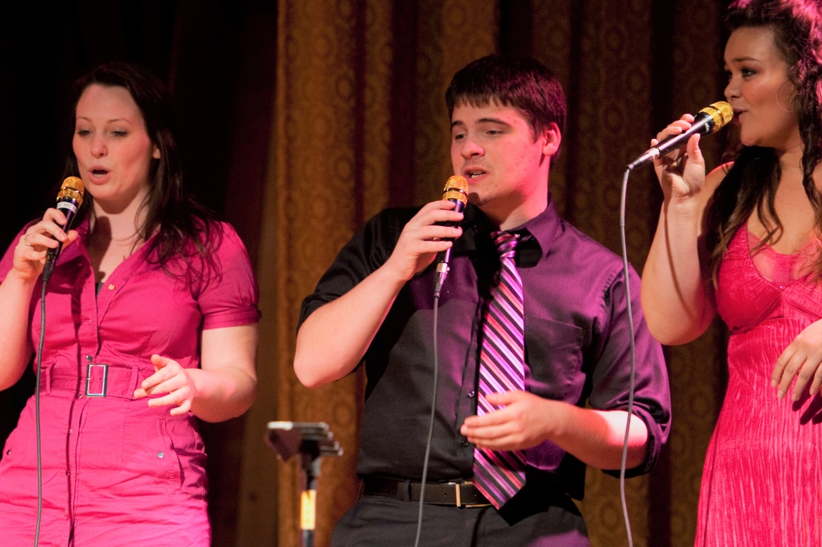 Students on stage under lights wearing bright pink and black singing into microphones with expressive faces