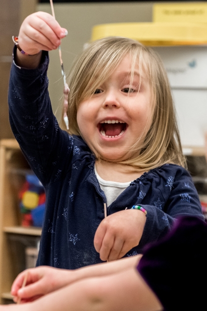 Little sandy haired girl sitting at table with a giant happy smile as she pulls on some gooey string.