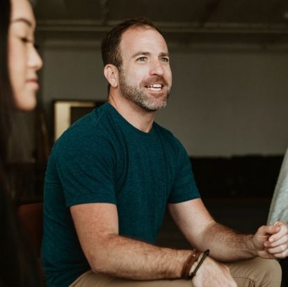 Man with short beard talks with others in a group setting, looking hopeful.