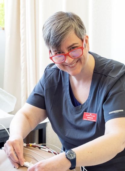 Mature female student wearing medical scrubs and peering over her glasses while smiling at the camera, places sensor lines onto a medical dummy laying on a gurney.