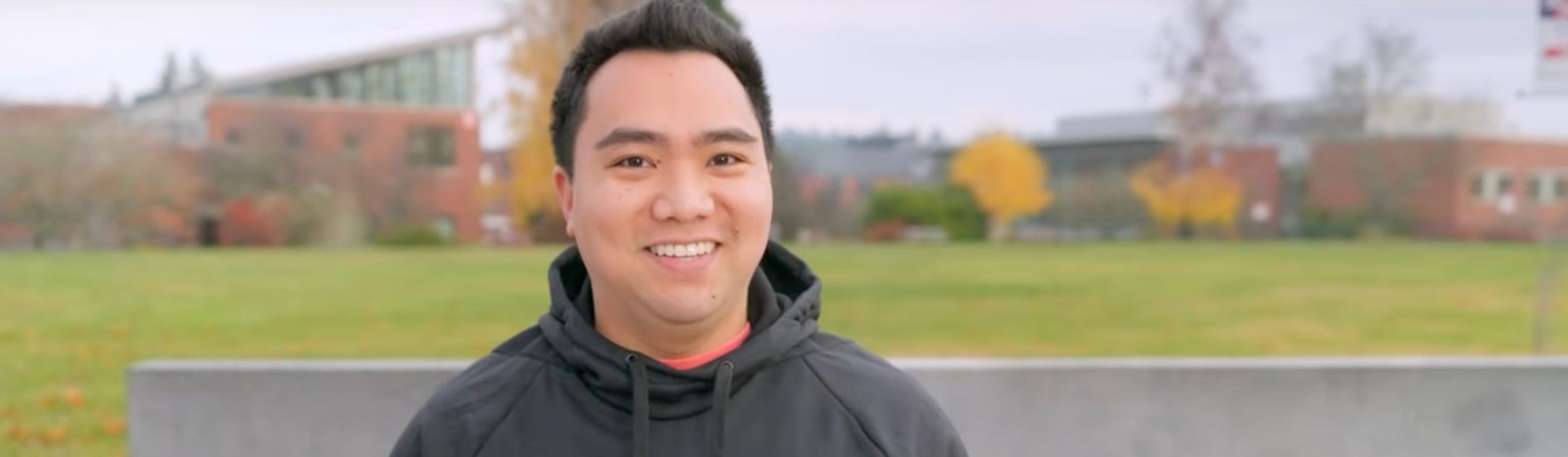 Employee in gray OC Rangers sweatshirt standing in front of concrete college sign.