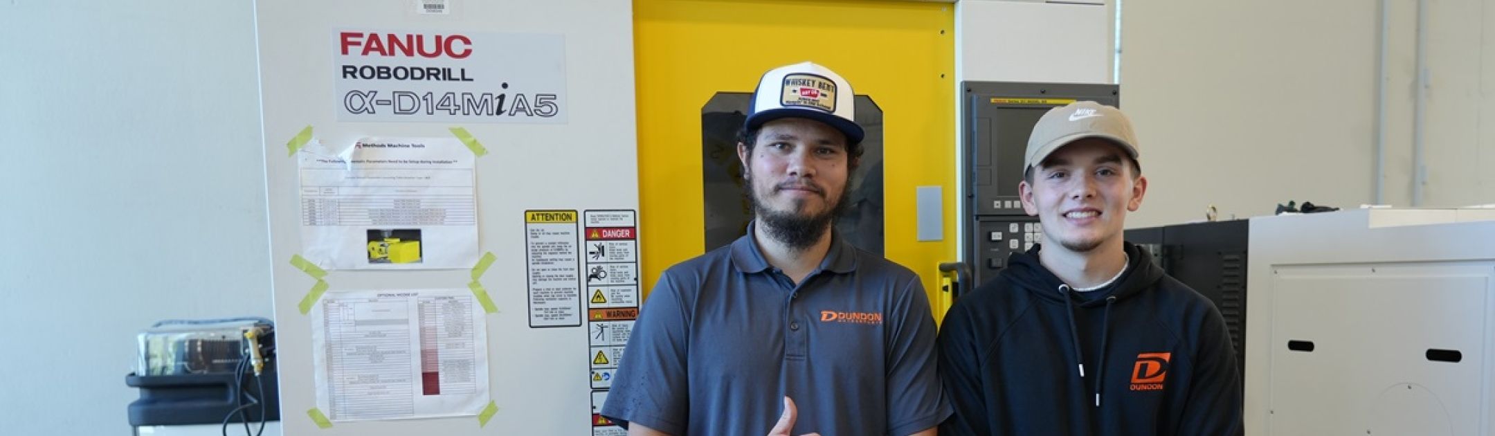 Welding students Justin Adams and Colin DeGross pose in front of a piece of equipment in the newly renovated Shops building