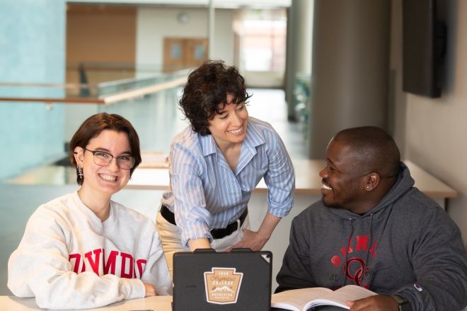 A faculty member with two students on either side of them, all looking at a tablet computer on the table in front of them.