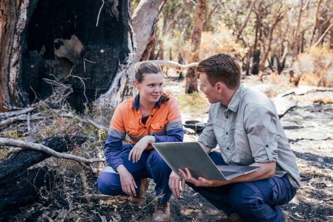 An instructor holding a laptop crouches with a student during a field study in the woods