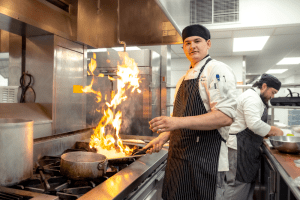 Smiling male student wearing kitchen cap and apron looks at the camera while holding a pan over the stove, a large flame jumping up out of the pan..