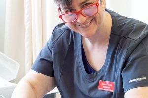 Mature female student wearing medical scrubs and peering over her glasses while smiling at the camera, places sensor lines onto a medical dummy laying on a gurney.