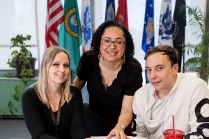Three people at table with flags in background. 
