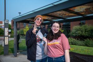 Two students in front of bus stop holding bus passes