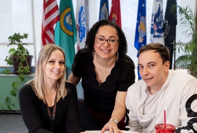 Three people at table with flags in background.