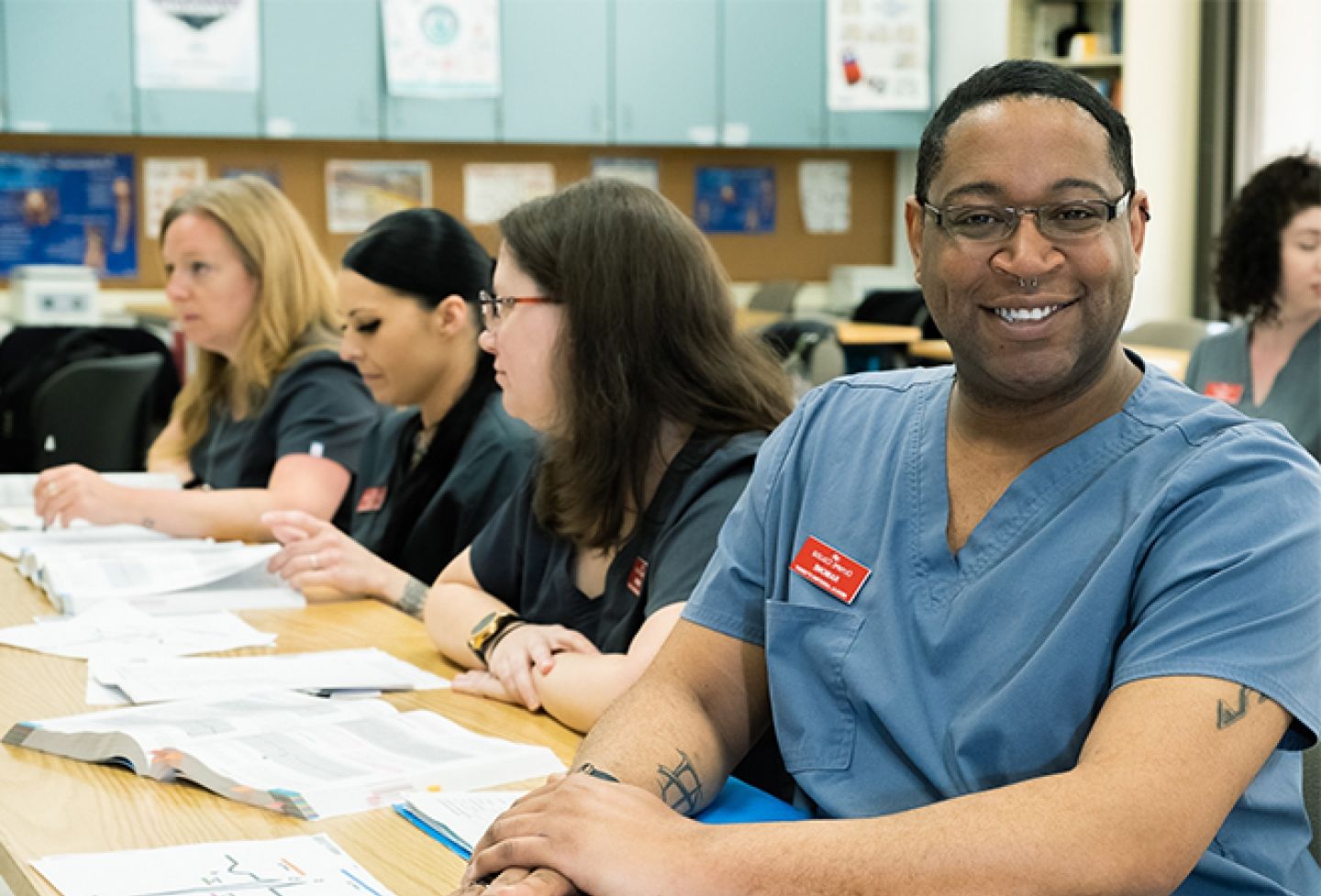 Male student in blue scrubs with classmates in background.