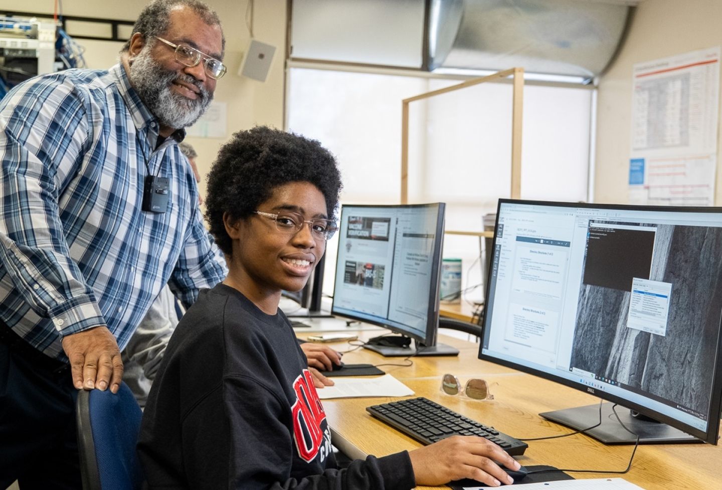 Student and instructor smiling at camera as they work at a computer station