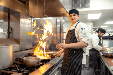 CulinaryHeader.jpg Smiling male student wearing kitchen cap and apron looks at the camera while holding a pan over the stove, a large flame jumping up out of the pan..