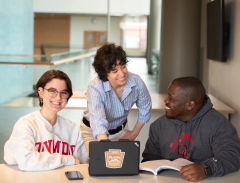 A faculty member with two students on either side of them, all looking at a tablet computer on the table in front of them.