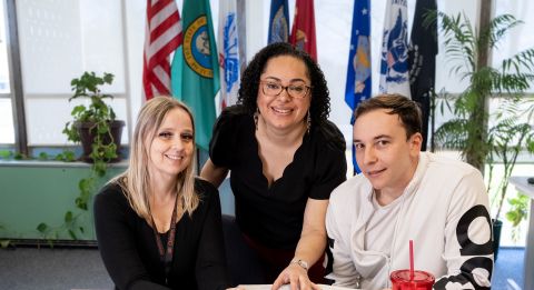 Three people at table with flags in background.