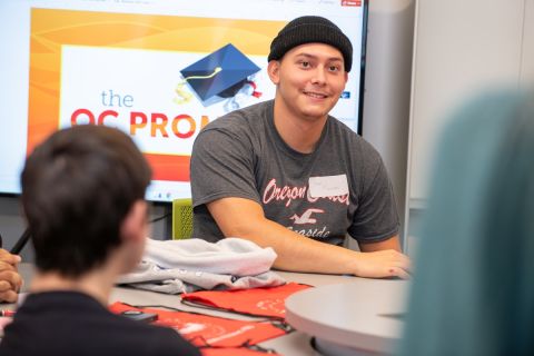 Student sitting in front of The OC Promise logo, with another student in the foreground looking on