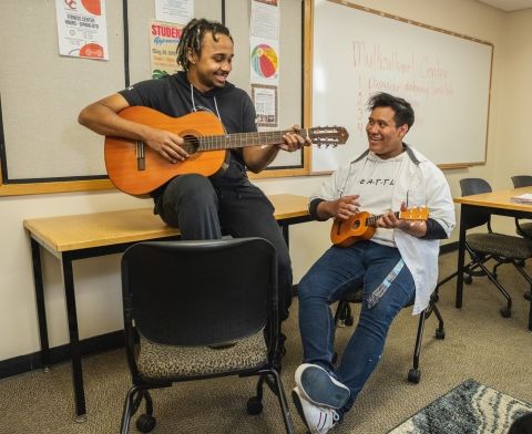 Two students in a classroom, one playing the guitar, the other playing a ukulele