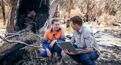 An instructor holding a laptop crouches with a student during a field study in the woods