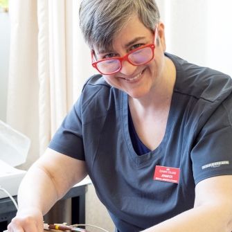 Mature female student wearing medical scrubs and peering over her glasses while smiling at the camera, places sensor lines onto a medical dummy laying on a gurney.