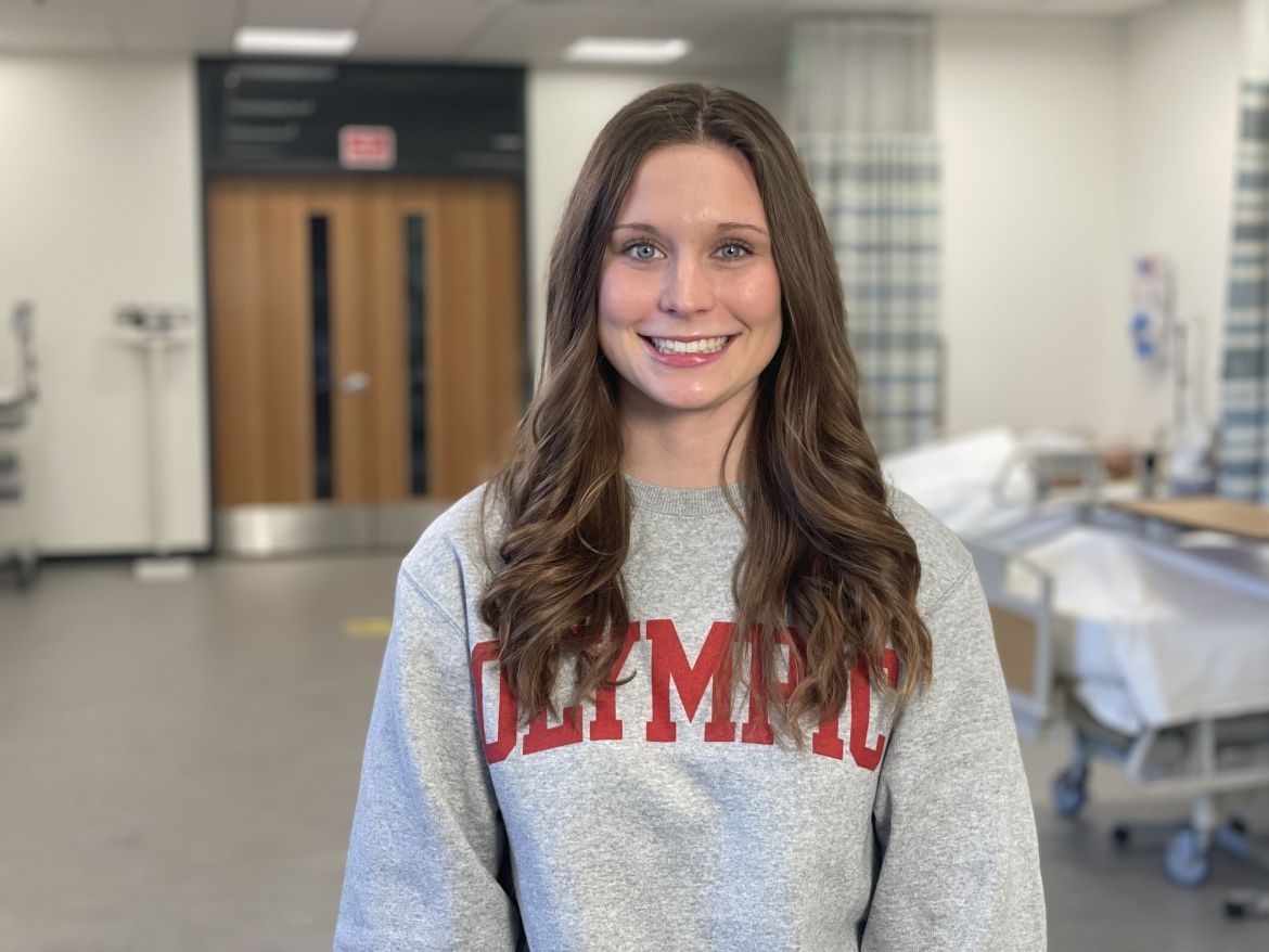 Smiling student wearing gray "Olympic" sweatshirt in nursing simulation lab