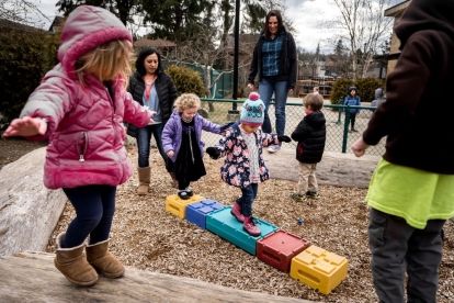 Children in winter coats play on a small playground with teachers in the mix.