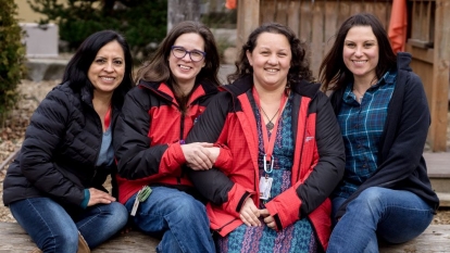 Group of four female teaching students with dark hair sit outside on a bench wearing winter coats, smiling at the camera.